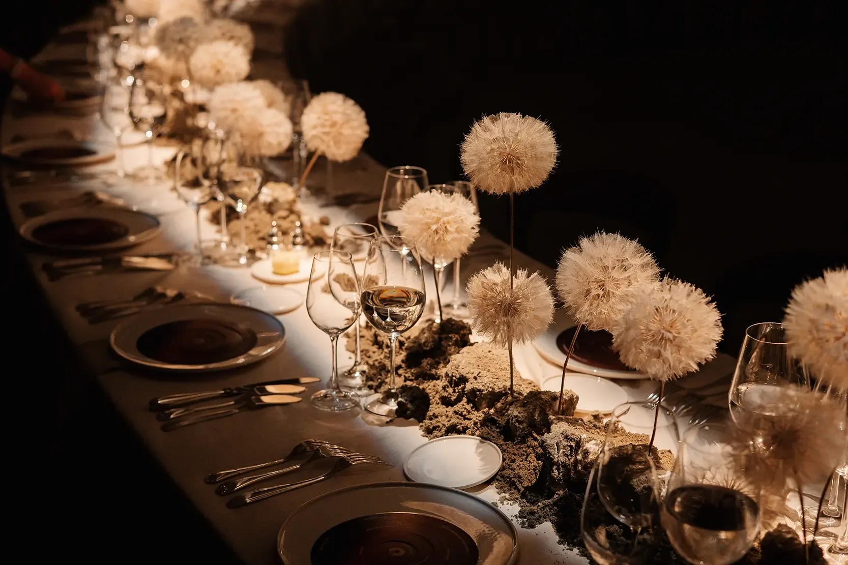 Lange gedekte tafel bij kaarslicht, met borden, glazen en gedroogde bolbloemen over de hele lengte.